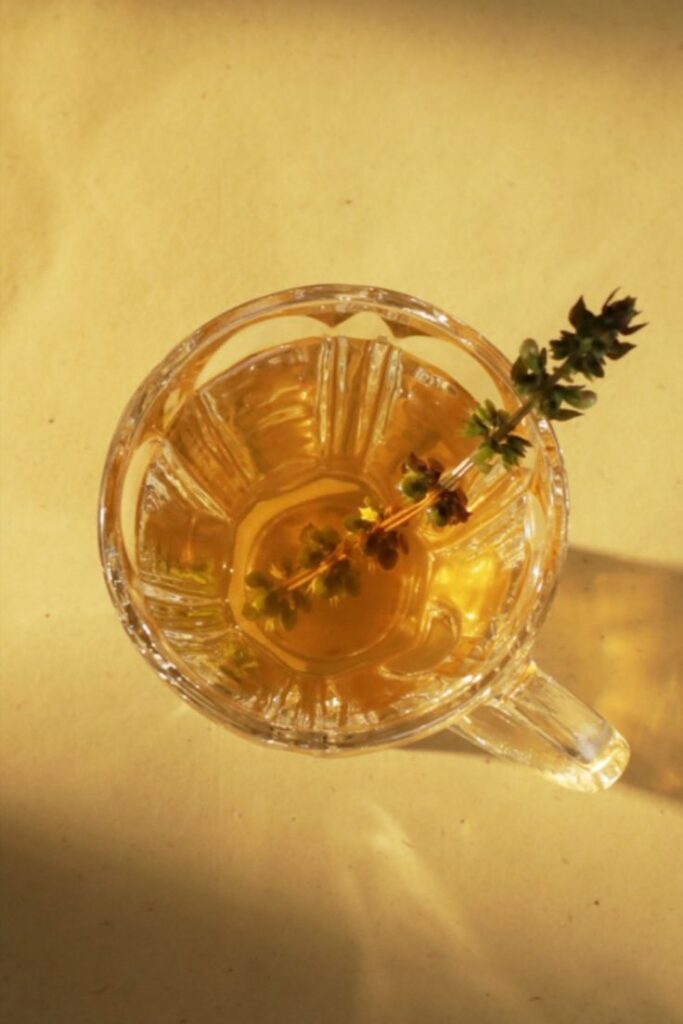 Clear glass of herbal tea with a sprig of fresh thyme on a sunny surface.