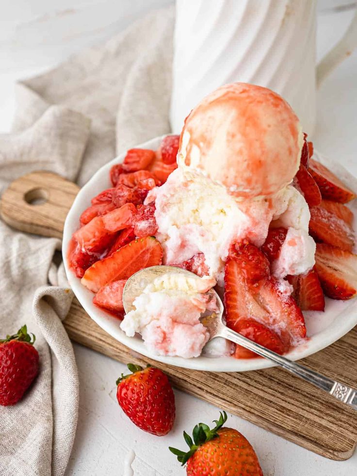 Bowl of vanilla ice cream topped with fresh strawberries and syrup, served with a spoon on a wooden board.
