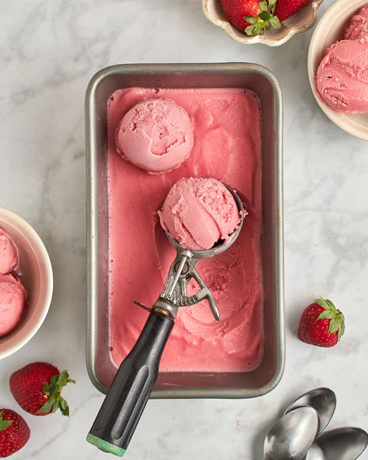 Strawberry ice cream in a metal tray with a scooper, surrounded by fresh strawberries and bowls.