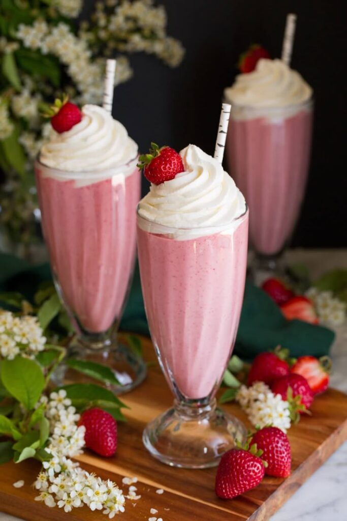 Three tall strawberry milkshakes with whipped cream and fresh strawberries on a wooden board.