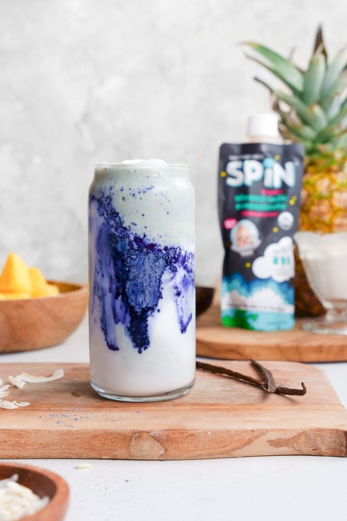 Creamy smoothie with blue swirl and fruit garnish on wooden board, next to pineapple and drink pouch.