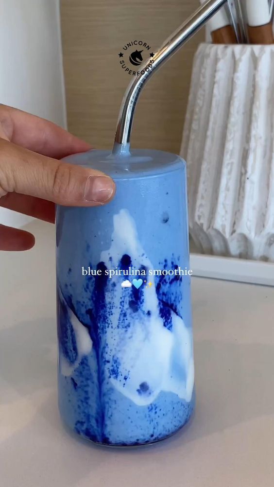 Blue spirulina smoothie in glass with metal straw, hand holding it; vibrant and nutritious drink.