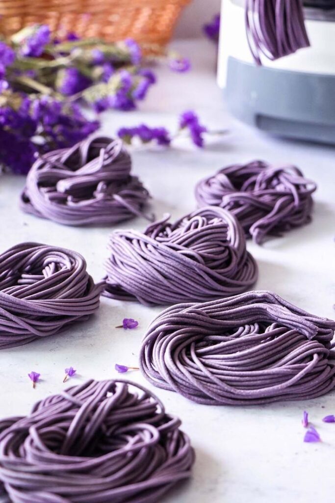 Purple pasta nests arranged on a white surface with purple flowers in the background.