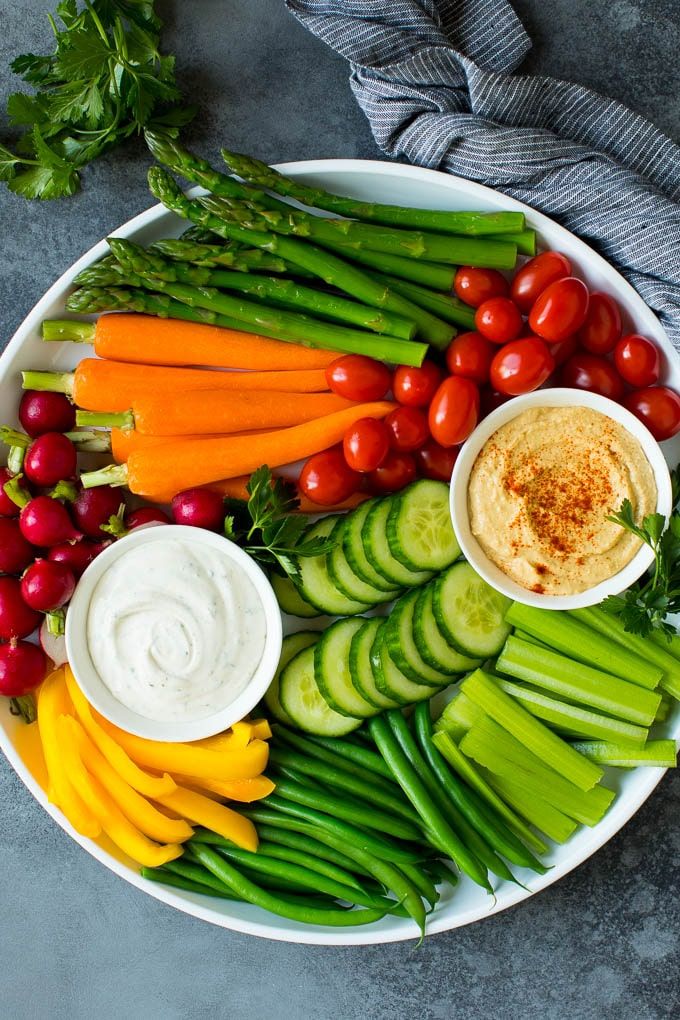 Colorful vegetable platter with dip, featuring carrots, asparagus, tomatoes, cucumbers, and more, served with hummus.