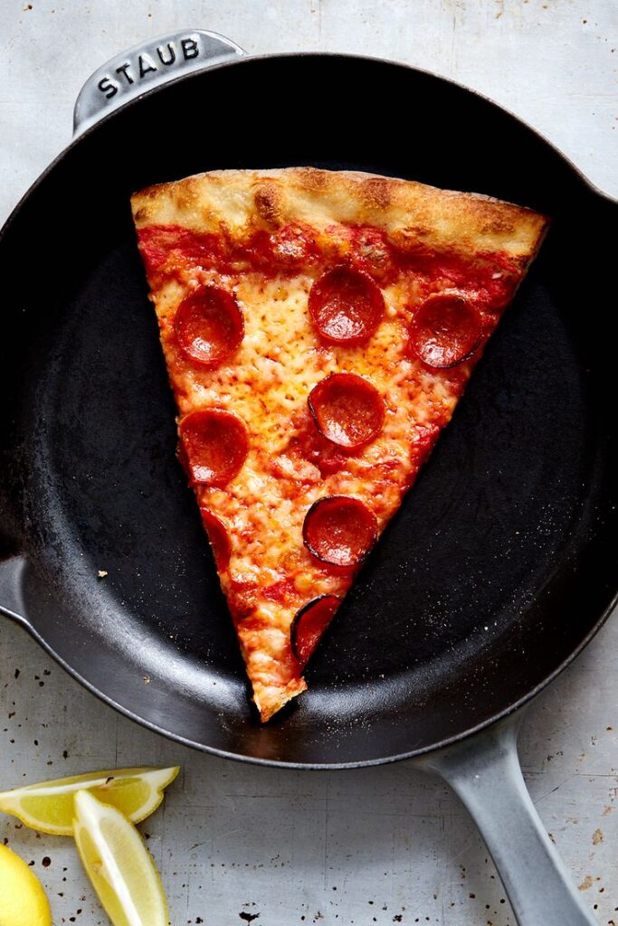 Slice of pepperoni pizza in a skillet with lemon wedges nearby, showcasing delicious toppings and crispy crust.