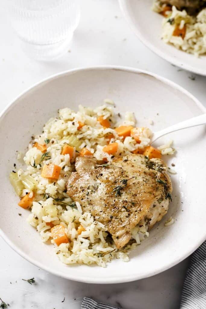 Herb-seasoned chicken thigh served with rice, carrots, and celery in a white bowl, garnished with herbs.