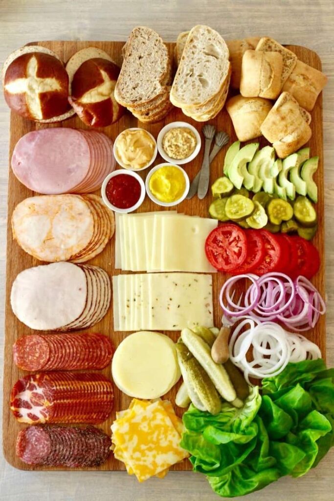 Charcuterie board with assorted breads, cheeses, meats, cucumbers, onions, pickles, tomatoes, and condiments.