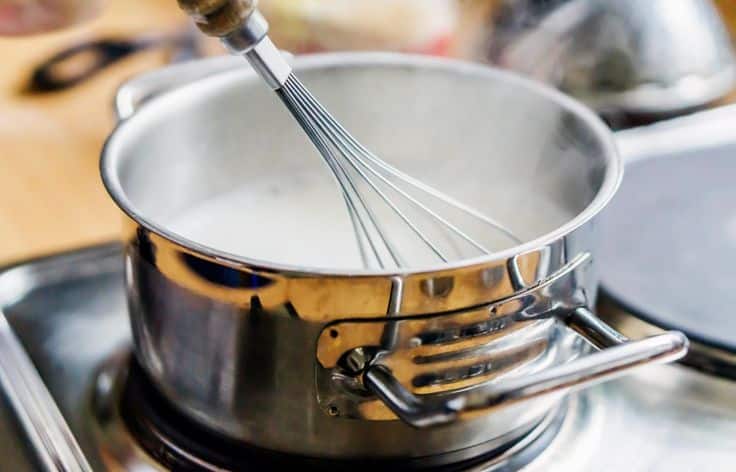 Whisk stirring creamy sauce in a stainless steel pot on the stove, steam rising, kitchen cooking scene.