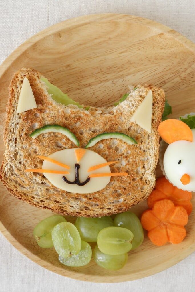 Cat-themed sandwich with cucumber and cheese decor, served with grapes and shaped eggs on a wooden plate.