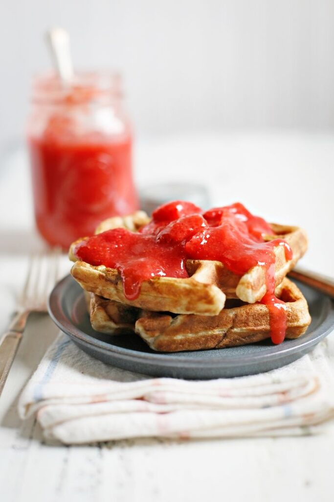 Waffles topped with fresh strawberry sauce on a plate, with jar in the background. Perfect breakfast treat.