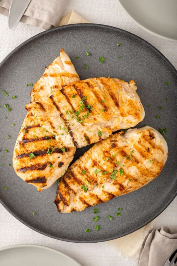 Grilled chicken breasts garnished with parsley on a gray plate, served on a light tablecloth with cutlery nearby.