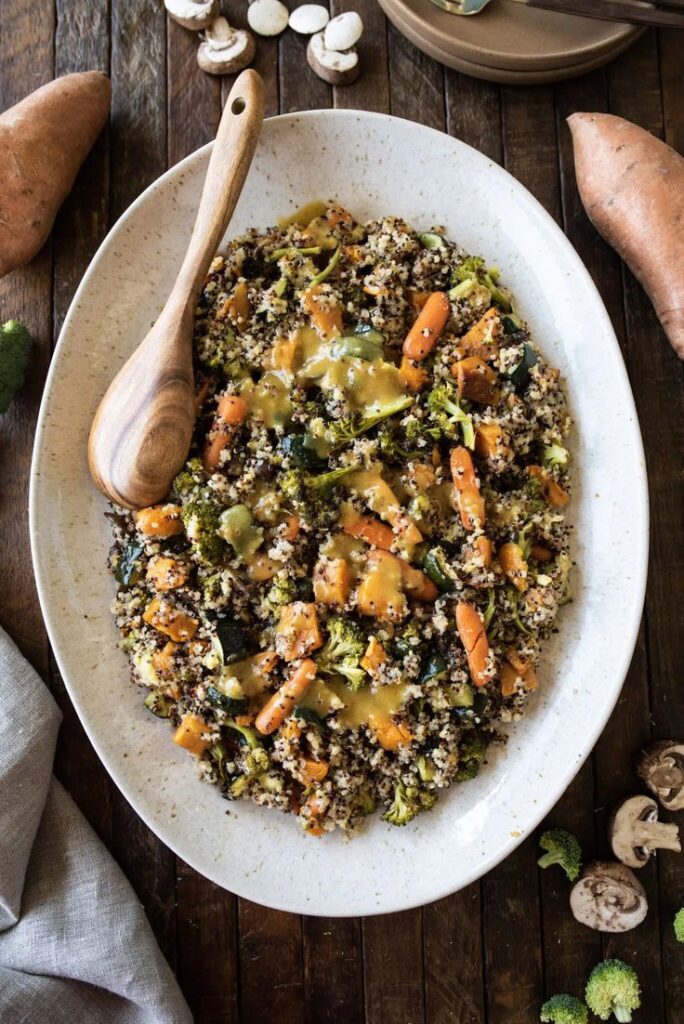 Quinoa salad with roasted vegetables and creamy dressing on a large white plate, garnished with broccoli and carrots.