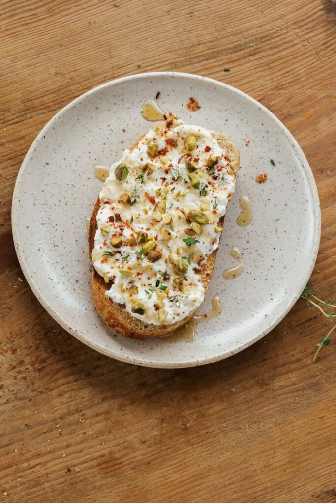Rustic toast topped with creamy cheese, pistachios, herbs, and honey, served on a speckled plate.
