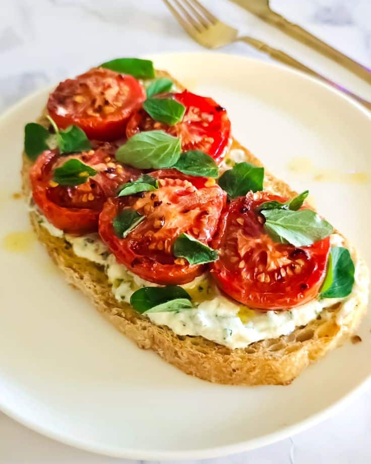 Open-faced sandwich with roasted tomatoes, fresh greens, and creamy spread on a white plate.