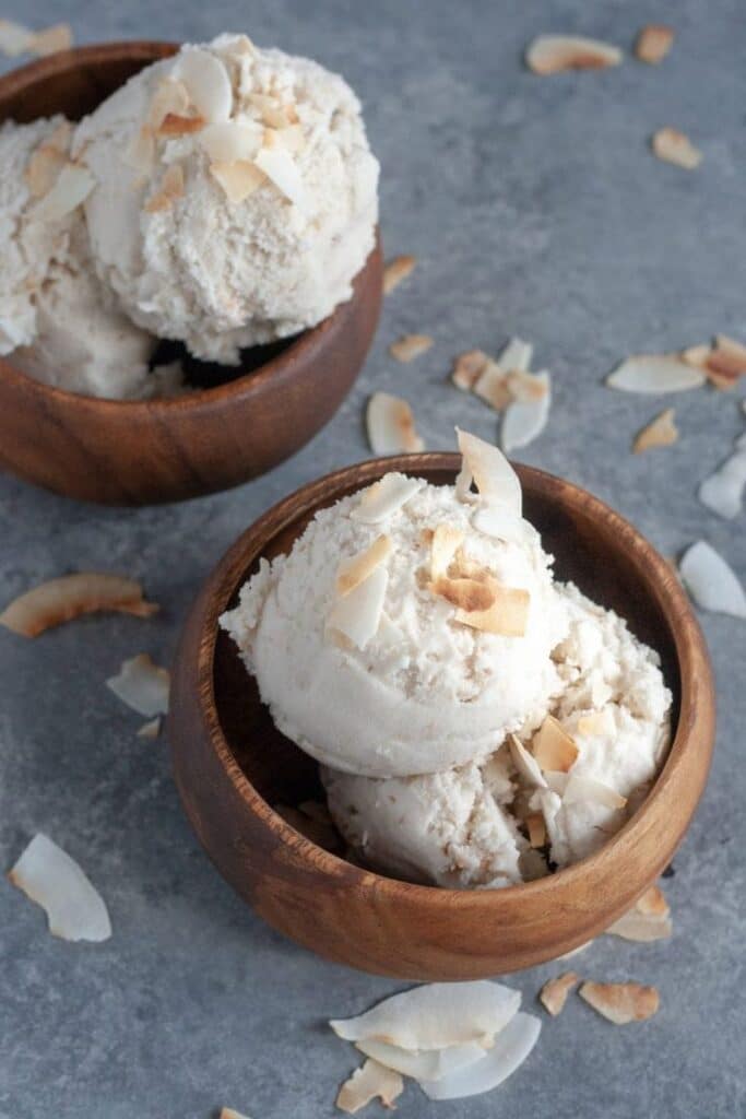 Two wooden bowls of coconut ice cream topped with toasted coconut flakes on a gray surface.