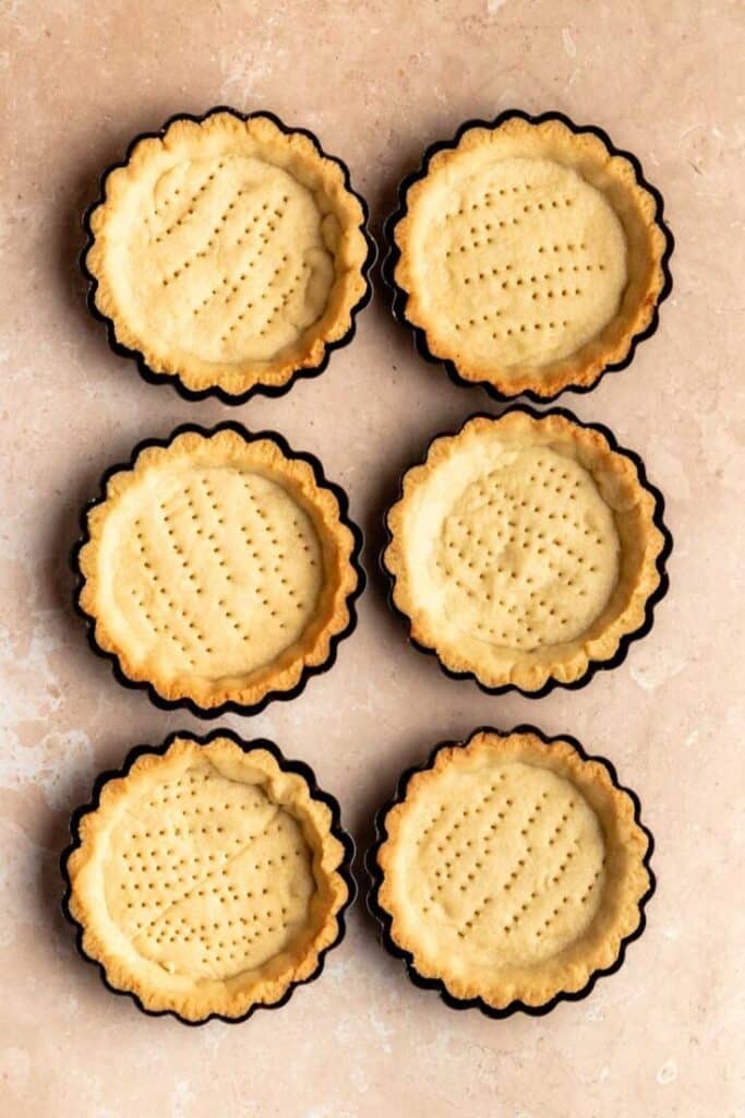 Six baked tart crusts on a beige surface, evenly arranged in two columns.