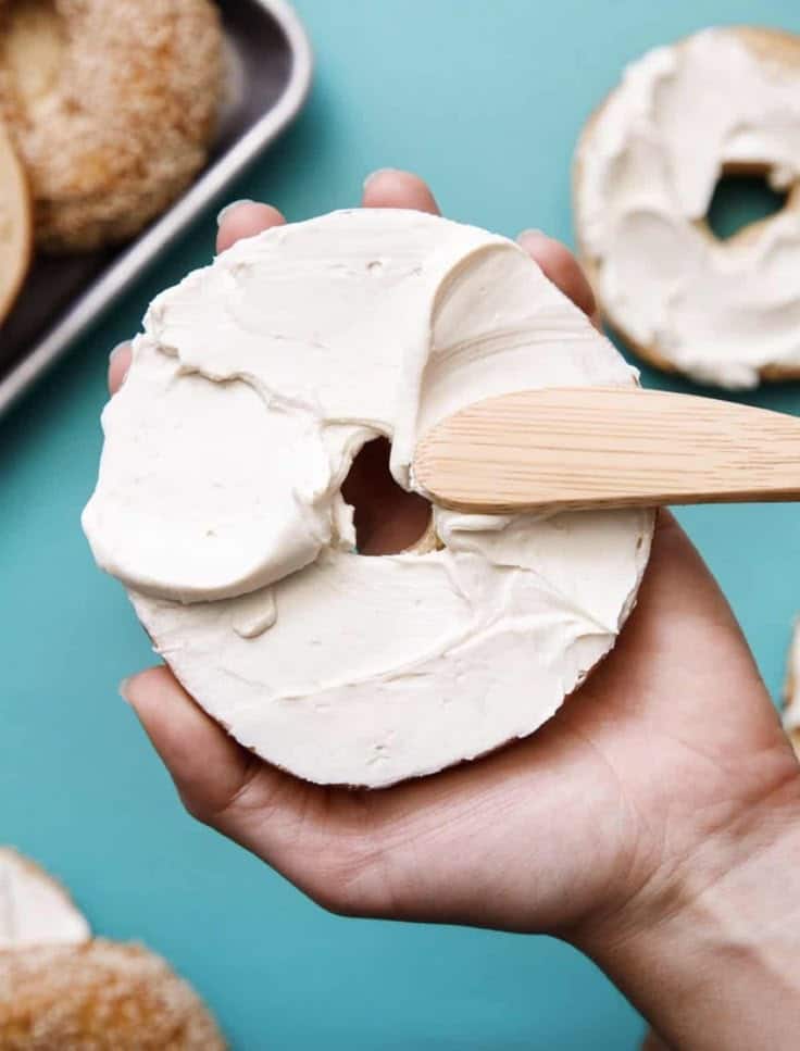 Spreading cream cheese on a bagel with a wooden spatula on a blue background.