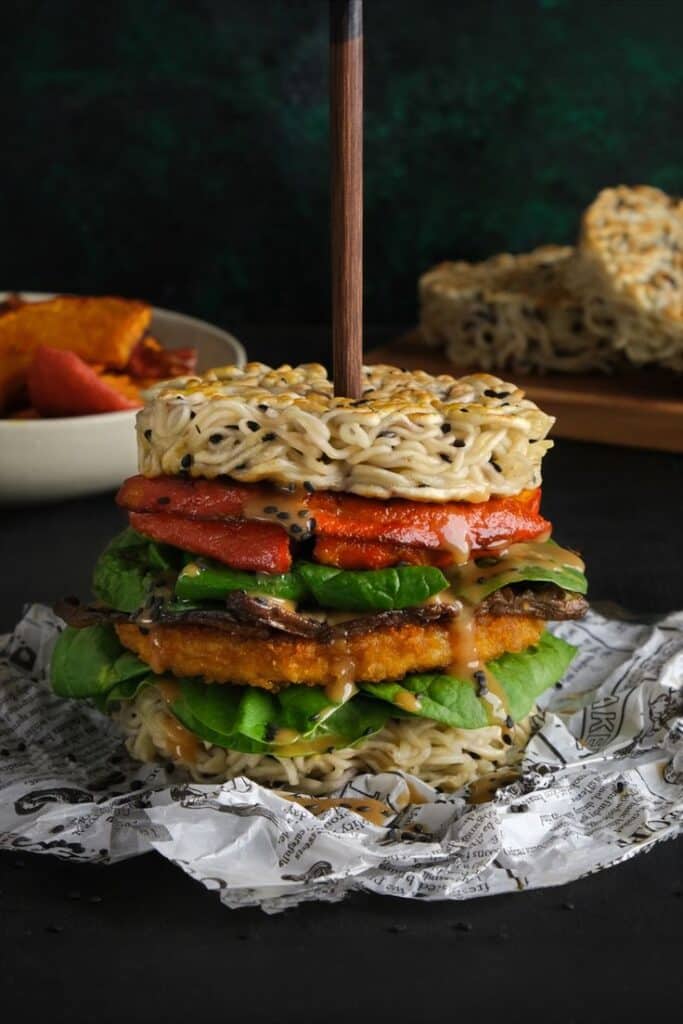 Gourmet ramen burger stacked with veggies and sauce, skewered, on a dark background with side dishes in view.