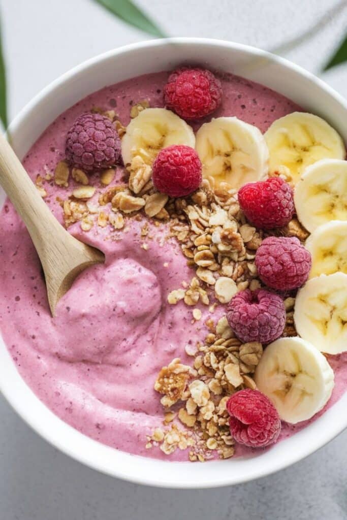 Smoothie bowl with raspberries, banana slices, and granola. Healthy breakfast with a wooden spoon.