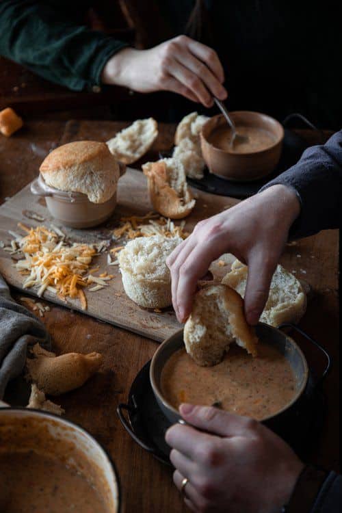 Hands dipping bread into creamy soup bowls on a rustic table with shredded cheese and bread rolls. Cozy dining scene.