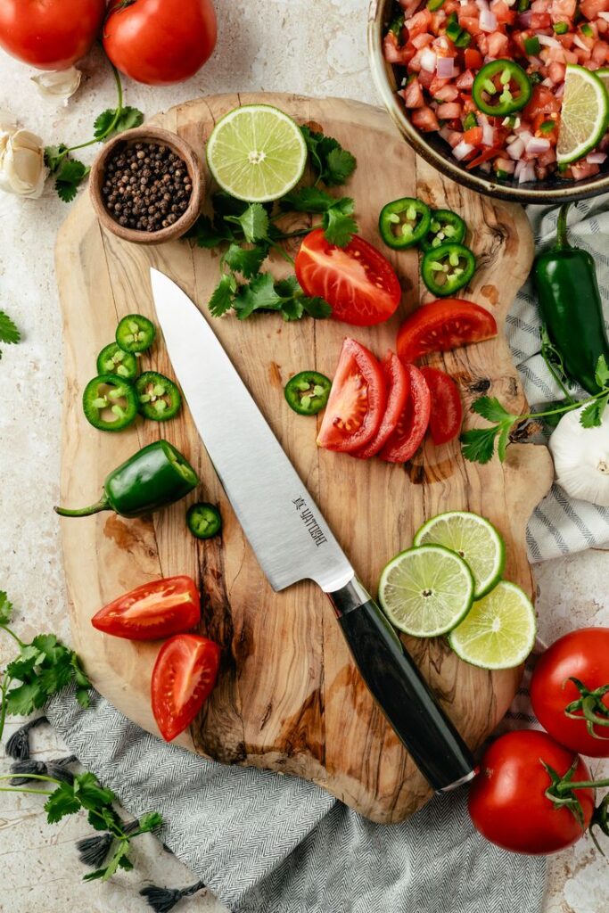 Fresh salsa ingredients on a wooden board with a knife—tomatoes, jalapeños, lime, and cilantro ready for chopping.