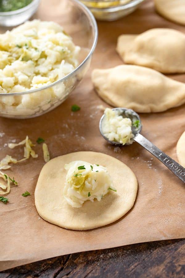 Dumpling preparation with potato filling on dough, surrounded by ingredients and measuring spoon on parchment paper.