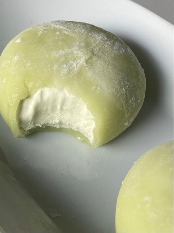 A close-up of a green mochi ice cream with a bite taken, revealing creamy filling on a white plate.