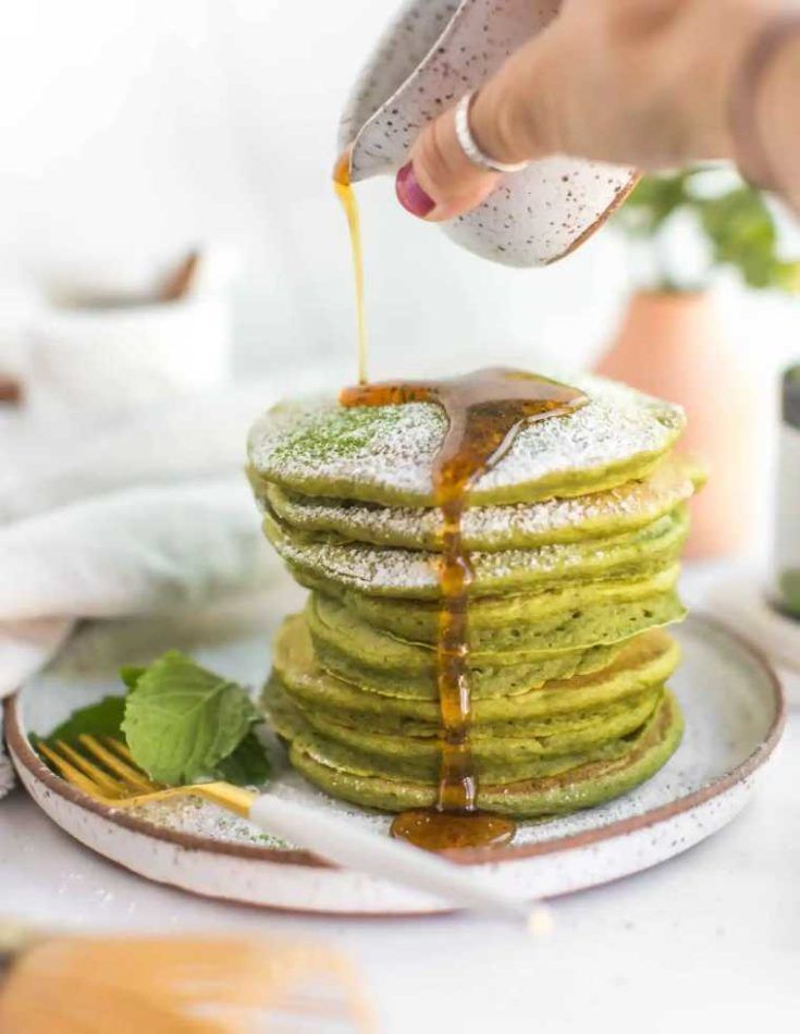 Stack of matcha pancakes with syrup poured on top, dusted with powdered sugar on a white plate.