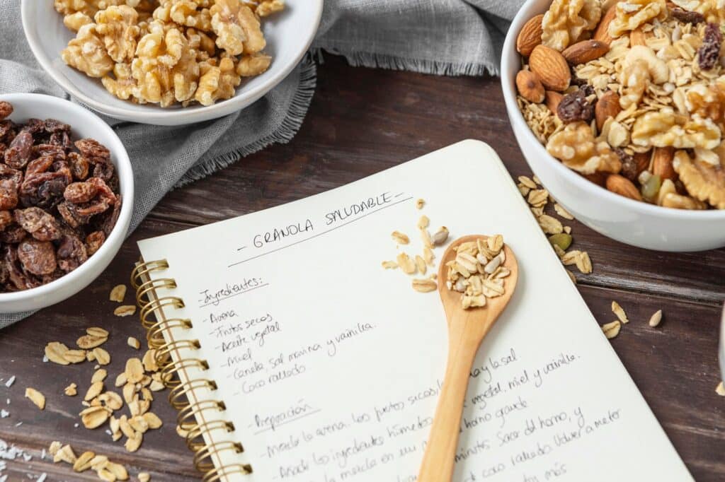Granola recipe with nuts and raisins, notebook and wooden spoon on a dark wooden table.