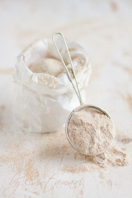 Sieve with whole wheat flour on a light surface, next to an open paper bag filled with more flour.