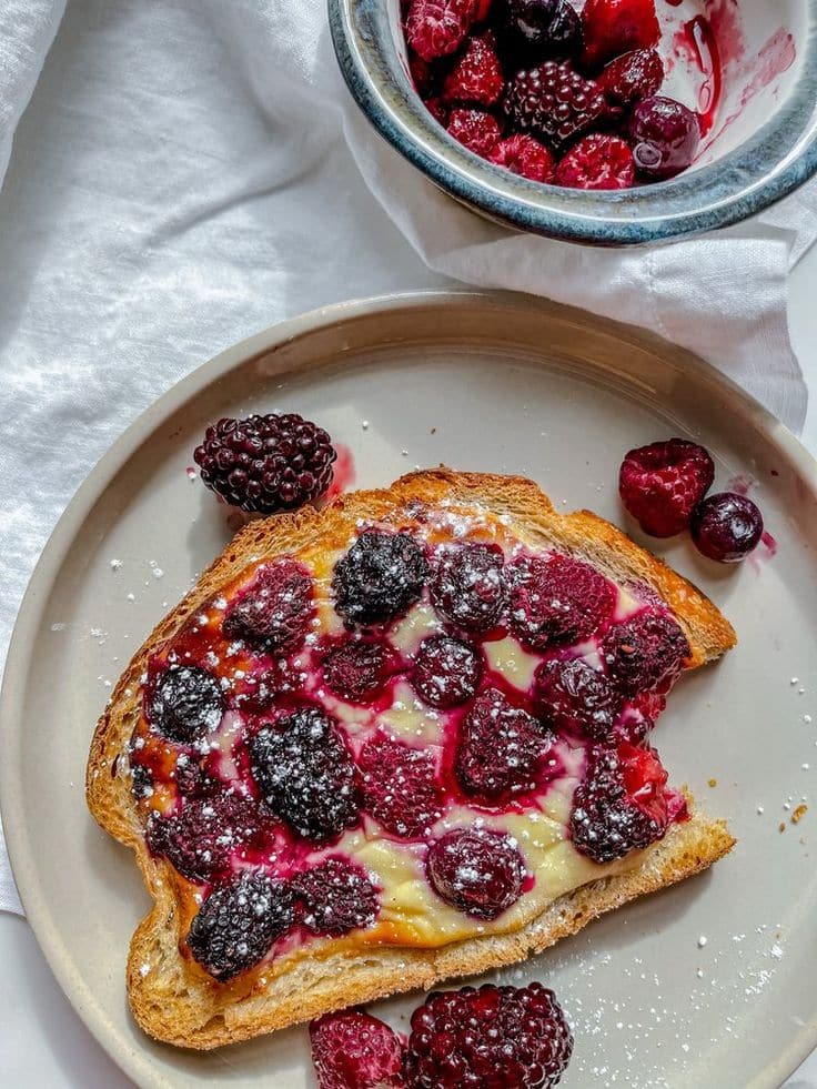 Toasted bread topped with melted cheese and mixed berries, served on a plate with fresh berries in a bowl nearby.