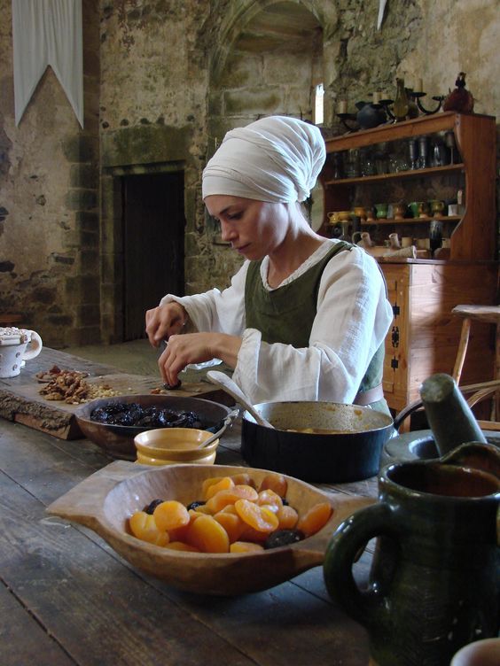 Medieval kitchen scene with a woman preparing dried fruits and nuts on a rustic wooden table.