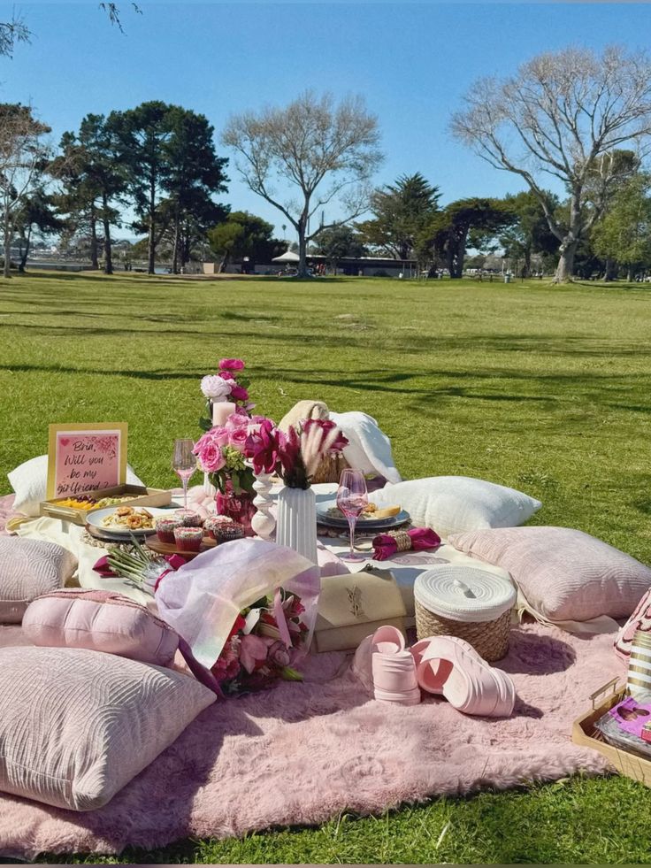 A cozy outdoor picnic setup on a sunny day with pink decor, flowers, and treats on a lush green lawn.