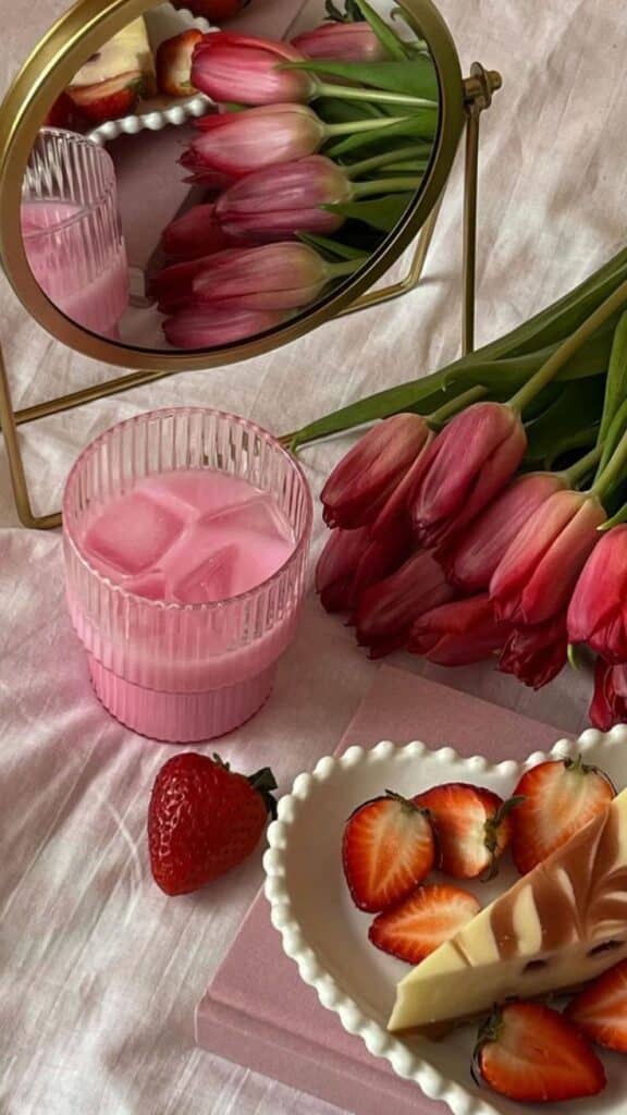 Reflective mirror with pink tulips, a glass of pink drink, strawberries, and a decorative plate on a soft pink background.