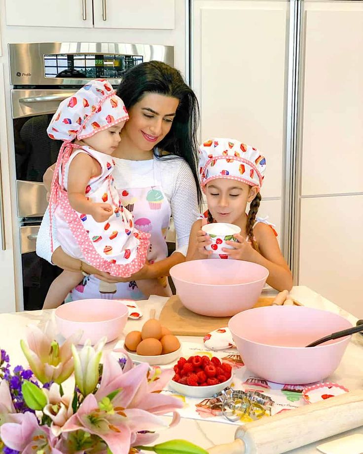 A mother and two children in a kitchen baking together, wearing matching aprons and hats, with ingredients around them.