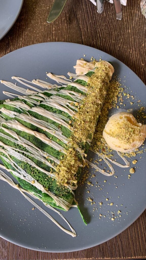 Green crepe with white drizzle and crushed peanuts, served with a scoop of ice cream on a gray plate.