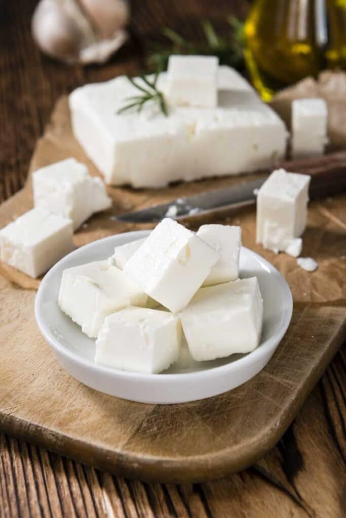 Cubes of fresh feta cheese on a wooden board with rosemary and olive oil in the background.