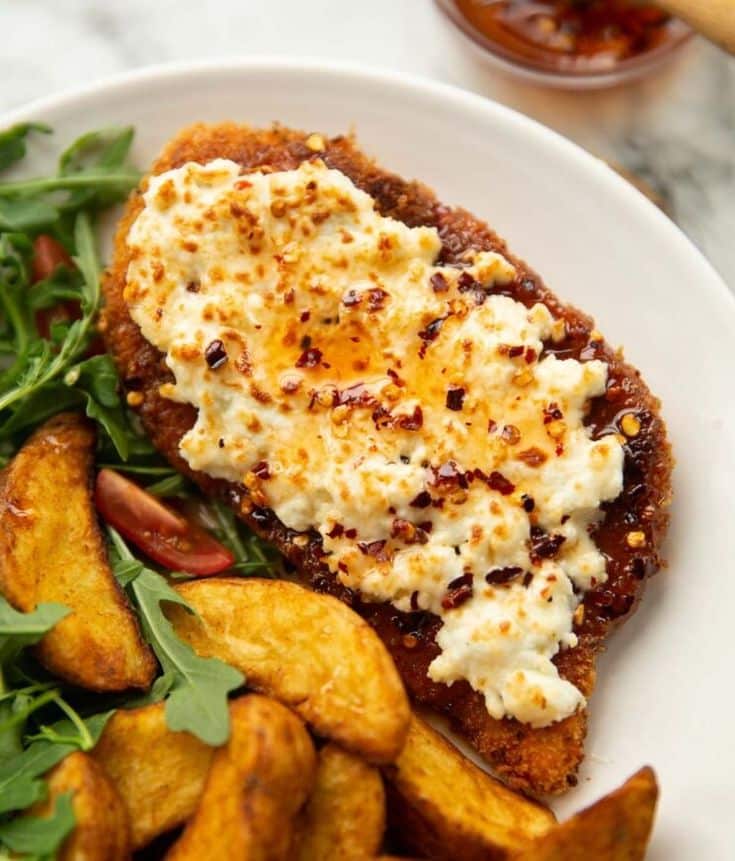 Crispy fried chicken with creamy topping, side of potato wedges, and fresh greens on a white plate.