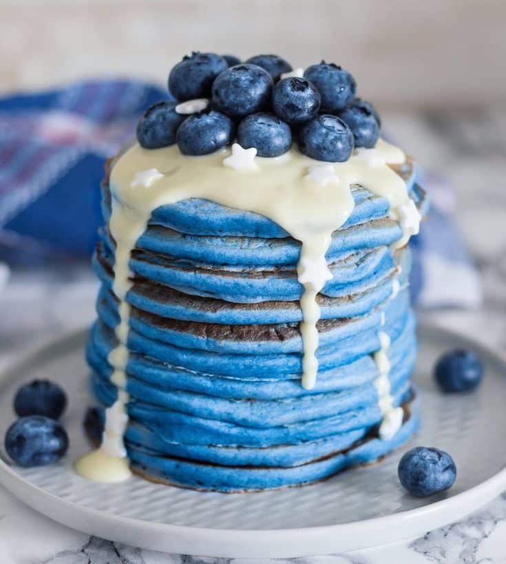 Stack of blue pancakes topped with fresh blueberries and drizzled with white syrup on a plate.