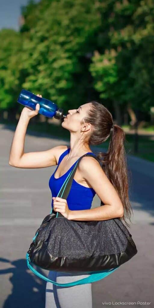 Woman in athletic wear drinking water outdoors, carrying a gym bag, emphasizing fitness and hydration.