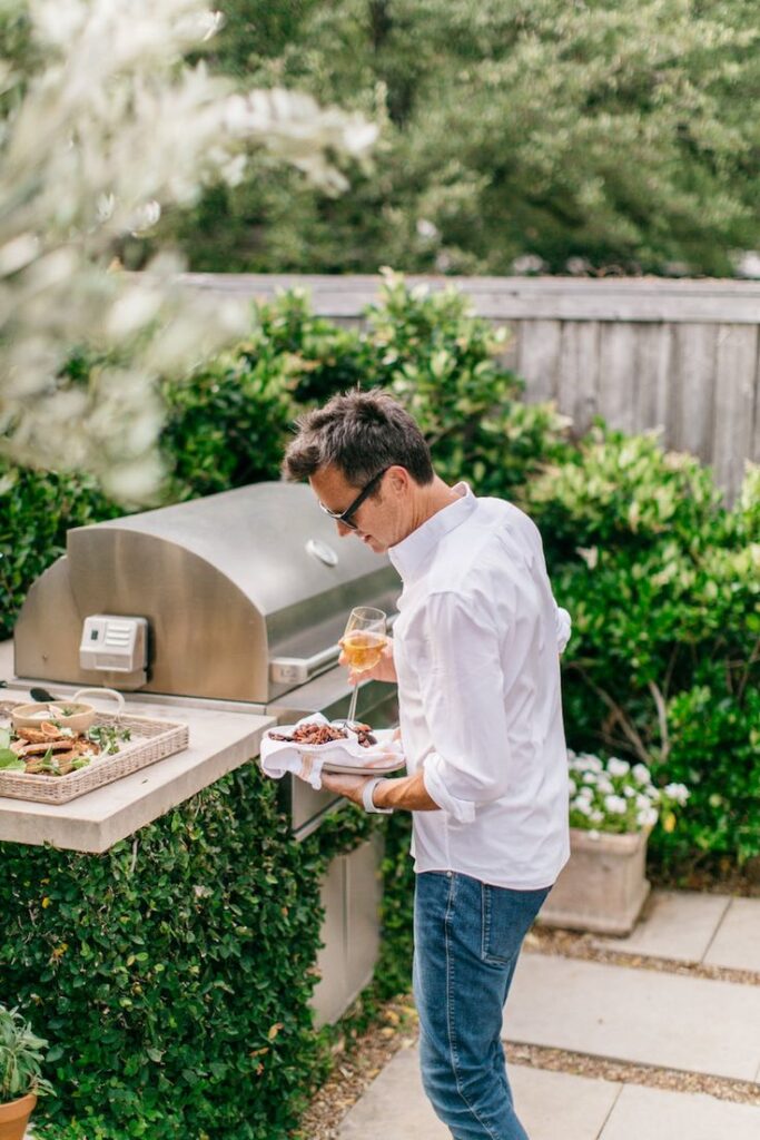 Man barbecuing outdoors with wine and food, enjoying a backyard cookout next to a grill surrounded by greenery.