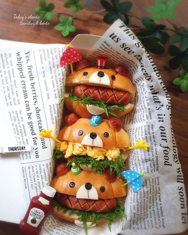 Adorable bear-shaped bento sandwiches with sausage, greens, and eggs, styled with playful picks and newspaper.
