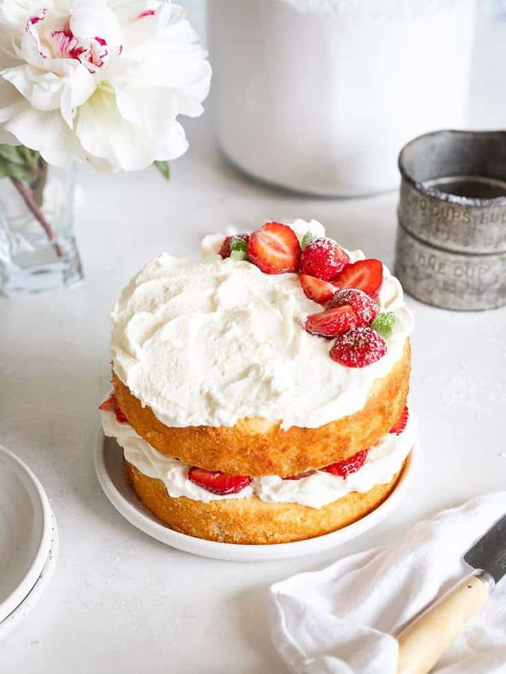 Two-layer vanilla cake with whipped cream and fresh strawberries, next to a flower in a vase.