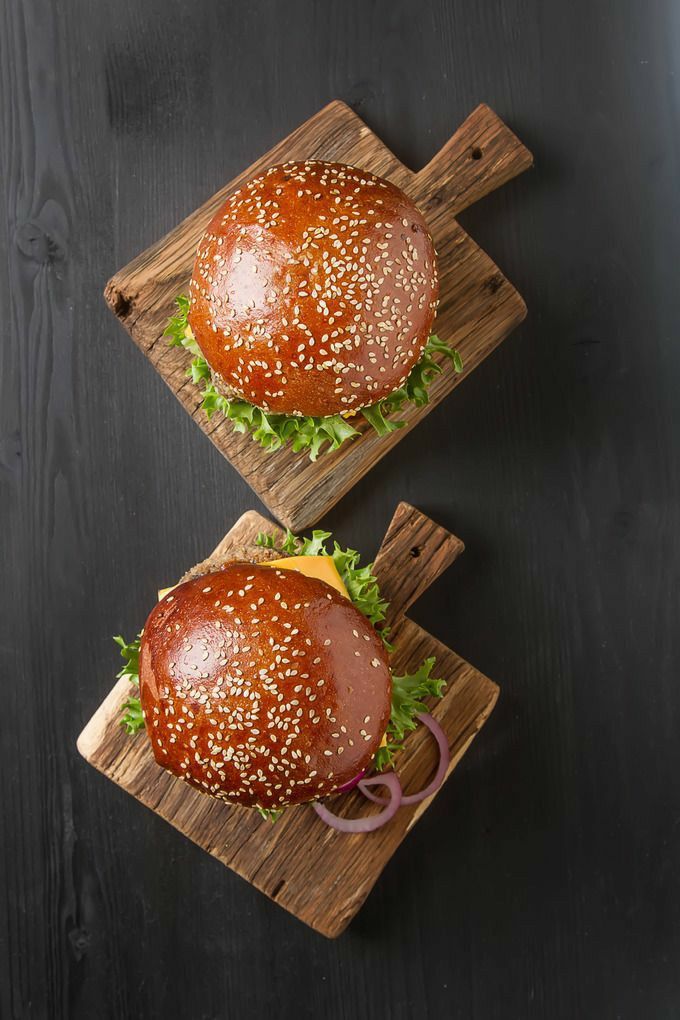 Two gourmet burgers on wooden boards with sesame seed buns, fresh lettuce, and cheese, arranged on a dark tabletop.