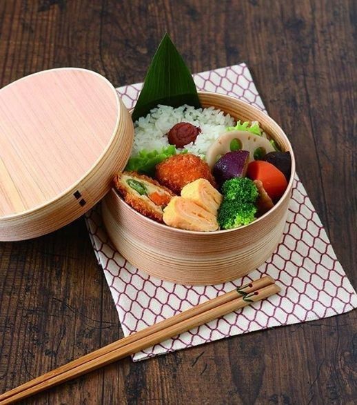 Wooden bento box with rice, vegetables, and rolled omelette on a wooden table, featuring traditional Japanese cuisine.