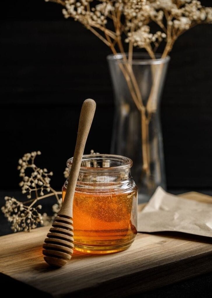 Jar of golden honey with a dipper on wooden board, dried flowers in vase, rustic and natural setting.