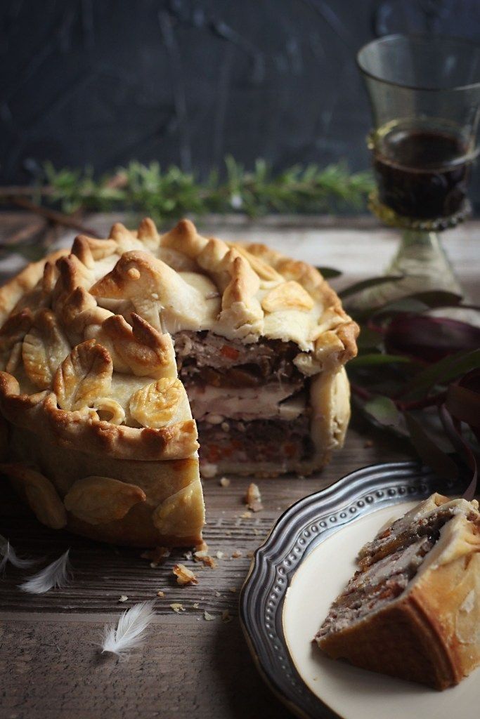 Rustic meat pie with detailed crust, sliced to reveal layers of filling, served on a wooden table with a drink.
