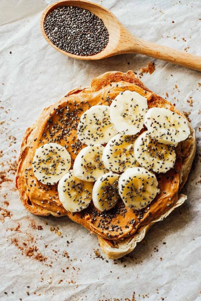 Toasted bread with peanut butter, banana slices, and chia seeds on parchment paper with a spoon of chia seeds.