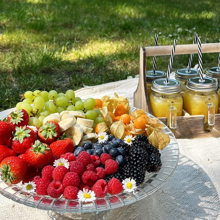 A colorful fruit platter with berries, grapes, and more, next to mason jars with juices, set on a picnic blanket.