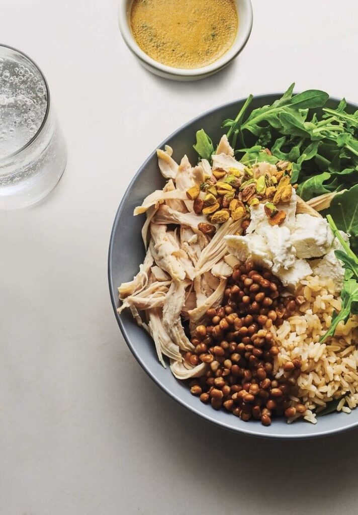 Healthy bowl with shredded chicken, chickpeas, rice, arugula, pistachios, and dressing. Glass of water on the side.